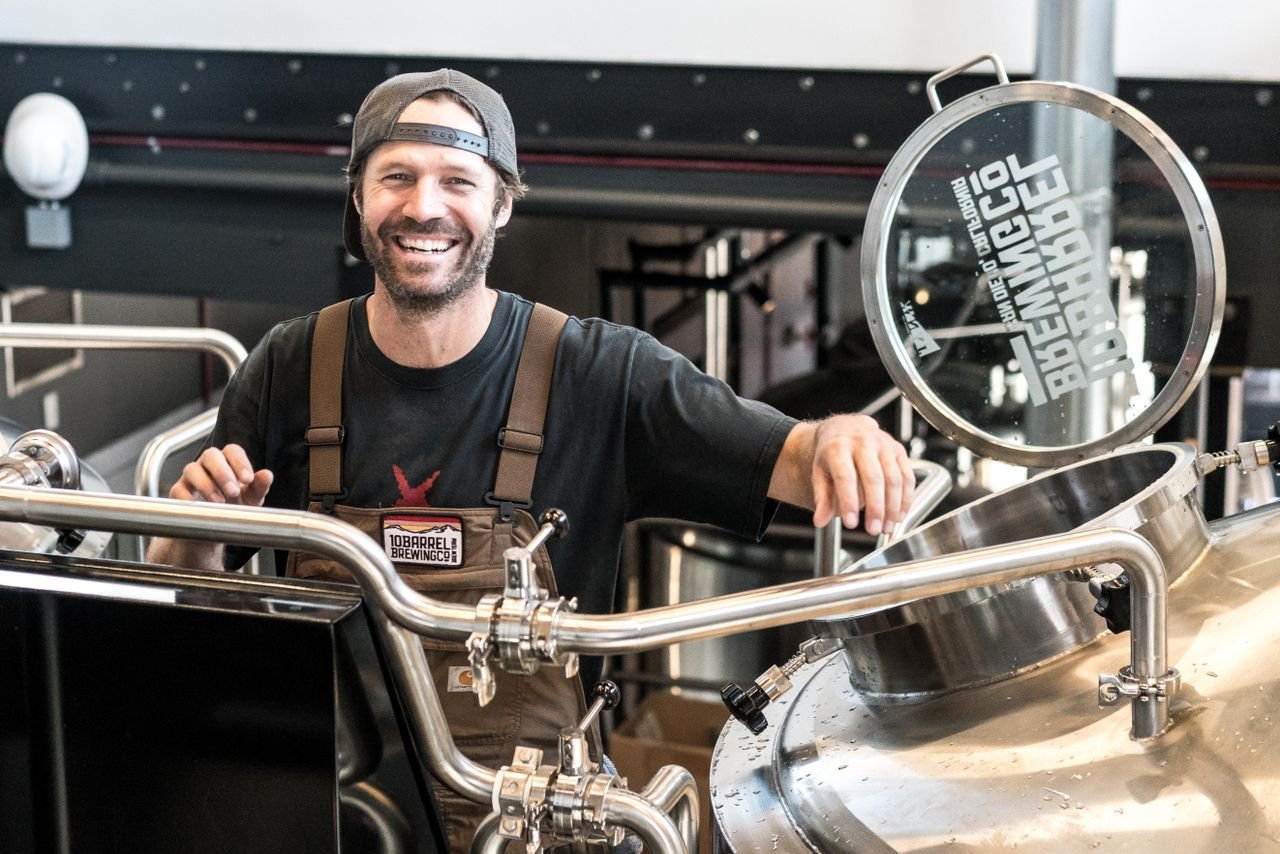 Smiling homebrewer lifting a large grain bag from a brew kettle with steam rising around them